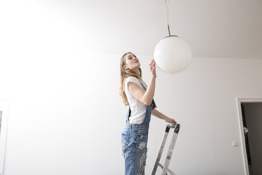 Junge Frau steht auf einer Leiter und prüft die Deckenleuchte - FMKF002513
