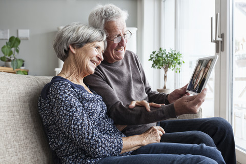 Älteres Ehepaar, das in seinem Wohnzimmer mit einem digitalen Tablet sitzt und mit seiner Familie skyped, lizenzfreies Stockfoto
