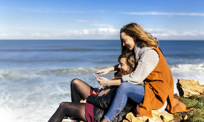 Spanien, Gijon, zwei junge Frauen haben Spaß mit einem Smartphone in der Nähe des Meeres - MGOF001496