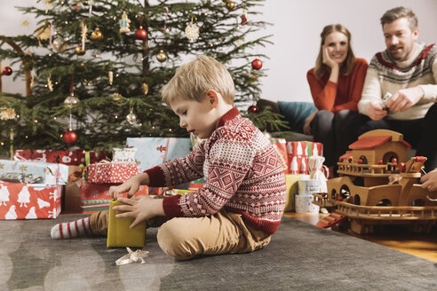 Kleiner Junge öffnet Weihnachtsgeschenk mit Eltern im Hintergrund - MFF002752
