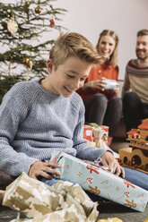 Little boy unwrapping a Christmas gift, parents sitting on couch in background - MFF002757