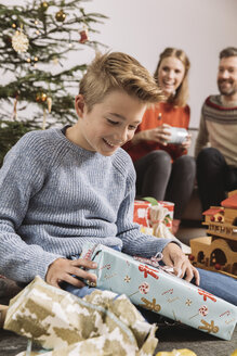 Kleiner Junge packt ein Weihnachtsgeschenk aus, die Eltern sitzen auf der Couch im Hintergrund - MFF002757