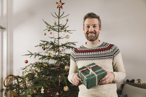 Porträt eines Mannes mit Weihnachtsgeschenk vor einem Baum stehend - MFF002777