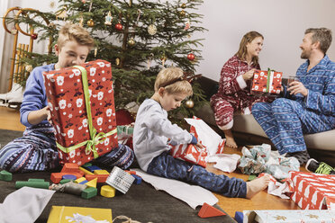 Kleiner Junge packt ein Weihnachtsgeschenk aus, die Eltern sitzen auf der Couch im Hintergrund - MFF002785