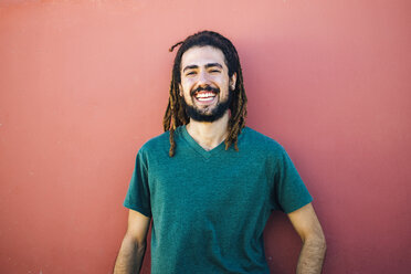 Portrait of laughing young man with dreadlocks and beard in front of a red wall - KIJF000232