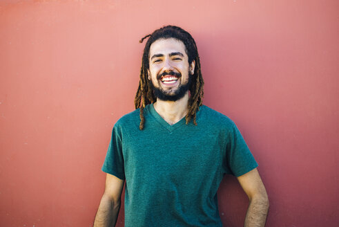 Porträt eines lachenden jungen Mannes mit Dreadlocks und Bart vor einer roten Wand - KIJF000232
