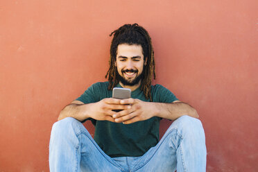 Porträt eines lächelnden jungen Mannes mit Dreadlocks und Bart, der vor einer rötlichen Wand auf sein Smartphone schaut - KIJF000234