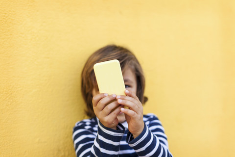 Kleiner Junge steht vor einer gelben Wand und spielt mit seinem Smartphone, lizenzfreies Stockfoto