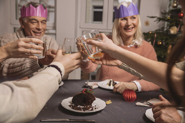 Happy senior couple with paper crowns clinking glasses while having Christmas pudding with their family - MFF002866