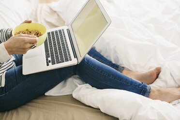 Frau auf dem Bett sitzend mit Laptop und Müslischale - EBSF001234