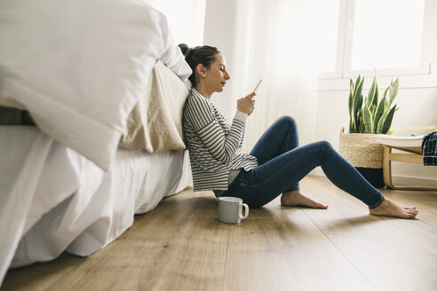Relaxed woman sitting in bedroom looking at cell phone - EBSF001239