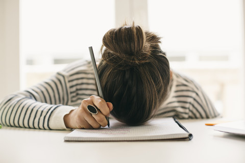 Frau schreibt auf Notizblock und stützt ihren Kopf auf den Tisch, lizenzfreies Stockfoto