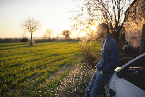 Mann lehnt an einem Auto und schaut auf eine grüne Wiese bei Sonnenuntergang - GEMF000785