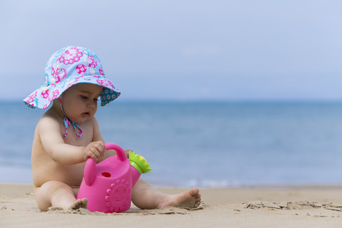 Kleines Babymädchen mit Sommerhut spielt am Strand mit Spielzeug Gießkanne, lizenzfreies Stockfoto