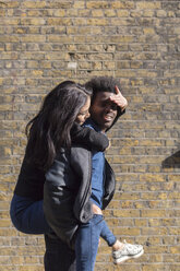 Young man carrying girlfriend piggyback at brick building - BOYF000202