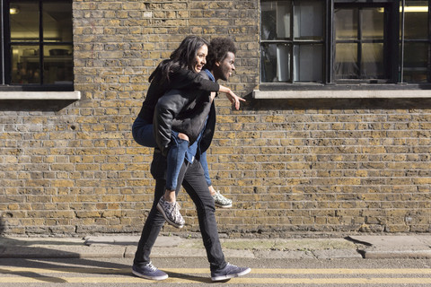 Junger Mann trägt Freundin huckepack an einem Backsteinbau, lizenzfreies Stockfoto
