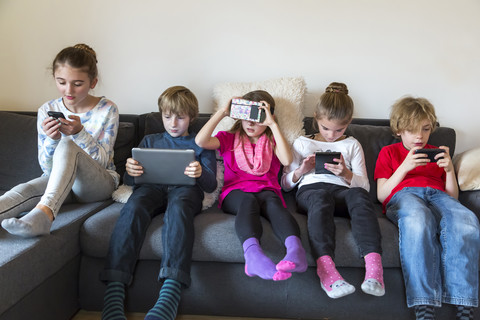 Gruppenbild von fünf Kindern, die auf einer Couch sitzen und verschiedene digitale Geräte benutzen, lizenzfreies Stockfoto