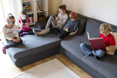 Vier Kinder auf einer Couch, die verschiedene digitale Geräte benutzen, während ein Junge ein Buch liest - SARF002644