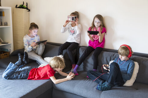 Fünf Kinder auf einer Couch, die verschiedene digitale Geräte benutzen - SARF002645