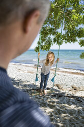 Smiling girl standing on a swing on the beach looking at her father in the foreground - OJF000125