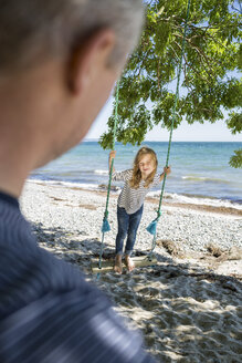 Lächelndes Mädchen, das auf einer Schaukel am Strand steht und ihren Vater im Vordergrund betrachtet - OJF000125