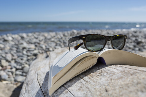 Sonnenbrille auf aufgeschlagenem Buch am Kieselstrand, Nahaufnahme - OJF000130