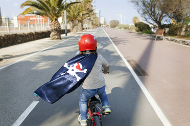 Spanien, Barcelona, Rückenansicht eines kleinen Jungen mit einem Piratenumhang, der auf einer leeren Straße Fahrrad fährt - VABF000368