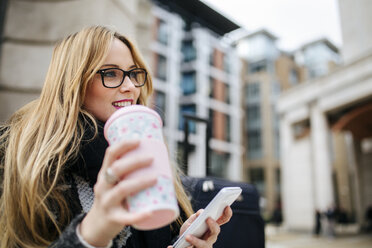 Lächelnde junge Frau mit Coffee to go und Mobiltelefon im Freien - MGOF001534