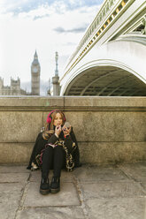 UK, London, young woman listening music and painting her lips near Westminster Bridge - MGOF001554