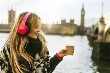 UK, London, young woman listening music and drinking coffee near Westminster Bridge - MGOF001556