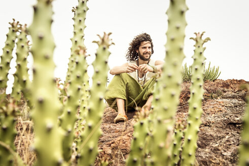 Man sitting on rock behind cacti, smiling - BMAF000107