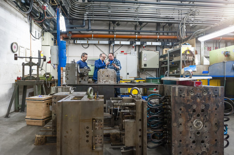 Drei Männer in einer Fabrik sprechen über ein Metallwerkstück, lizenzfreies Stockfoto