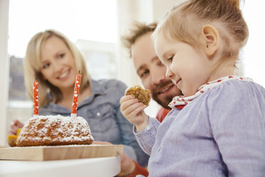 Kleines Mädchen isst ein Stück von ihrem Karotten-Geburtstagskuchen, während die Eltern zusehen - MFF002883