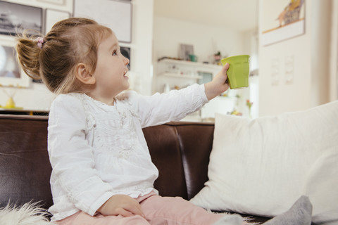 Kleines Mädchen hält einen Plastikbecher hoch und sitzt auf der Couch, lizenzfreies Stockfoto