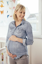 Porträt einer schwangeren Frau, die ihren Bauch im Kinderzimmer hält - MFF002905