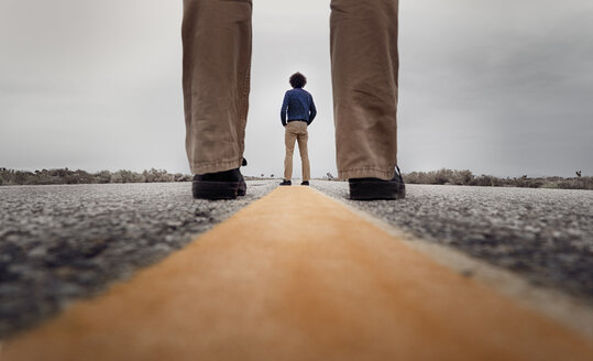 View through standing legs to a curly man standing on the medial strip of a road - BMA000202