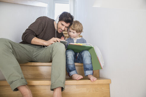 Vater und Sohn sitzen auf der Treppe, lesen ein Buch und hören Musik - FMKF002523