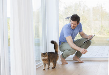 Mann mit digitalem Tablet, der eine Katze betrachtet - FMKF002583