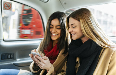 UK, London, Zwei junge Frauen benutzen ein Smartphone in einem schwarzen Taxi - MGOF001567