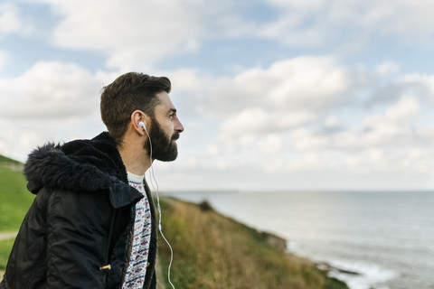 Profil eines bärtigen Mannes, der mit Kopfhörern Musik hört und auf das Meer schaut, lizenzfreies Stockfoto