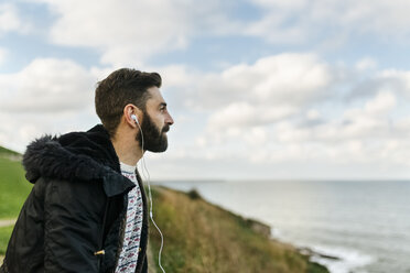 Profil eines bärtigen Mannes, der mit Kopfhörern Musik hört und auf das Meer schaut - MGOF001606