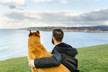 Spanien, Gijon, Rückenansicht eines Mannes, der mit seinem Hund auf einer Wiese sitzt und in die Ferne schaut - MGOF001608