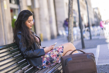 Young woman with baggage sitting on a bench looking at her smartphone - SIPF000272