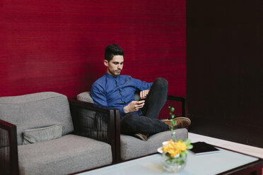 Junger Geschäftsmann sitzt auf einem Sessel in einer Lobby und benutzt sein Smartphone - BOYF000227