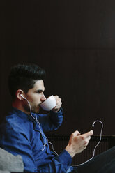 Profile of young man hearing music with earphones while drinking coffee in front of black background - BOYF000240