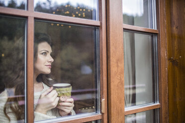 Young woman standing at window with cup of coffee - HAPF000320