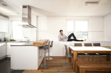 Mann liegt auf der Anrichte in seiner offenen Küche und benutzt sein Smartphone - MFRF000528