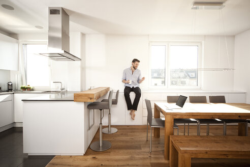 Mann mit Kaffeetasse, der auf der Anrichte in seiner offenen Küche sitzt und auf sein Smartphone schaut - MFRF000529