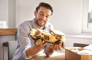Porträt eines glücklichen jungen Mannes, der am Tisch sitzt und ein hölzernes Spielzeugauto betrachtet - MFRF000537