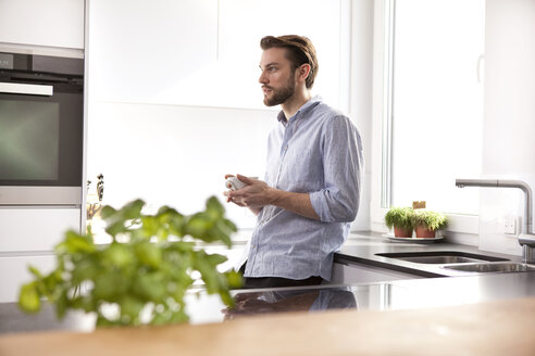 Nachdenklicher junger Mann mit einer Tasse Kaffee in seiner Küche stehend - MFRF000539